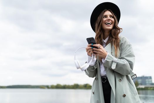 portrait of a smiling young woman in a black hat and autumn gray coat with a phone on the background of the water.