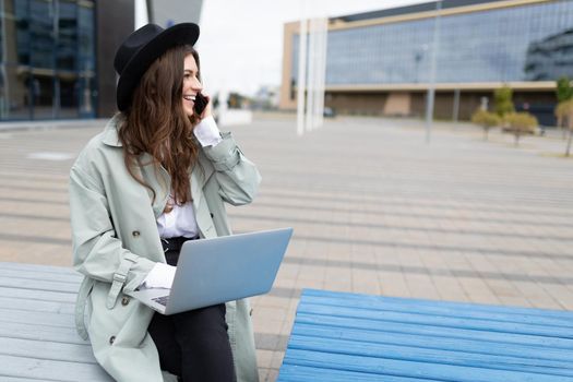 a young woman businesswoman works on a laptop and speaks on the phone well Sitting on a bench against the backdrop of an office building.