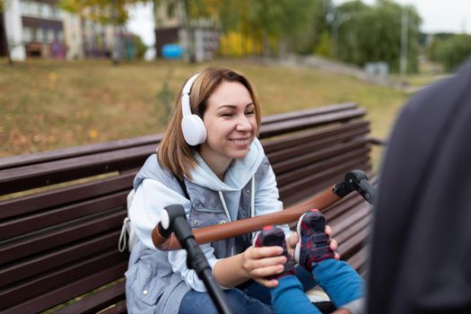 a young mother while walking with a small child in a stroller in headphones plays with him stroking his legs while sitting on a bench.