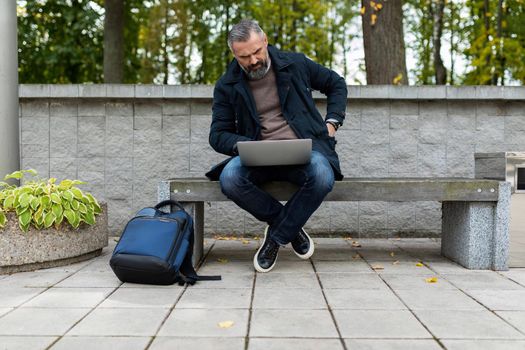 mature businessman man working online on a laptop sitting on a bench in a city park.
