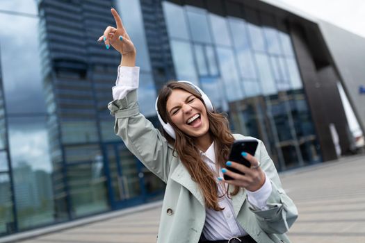 satisfied ambitious young woman dancing with a mobile phone in her hands and headphones against the backdrop of a glass office building.