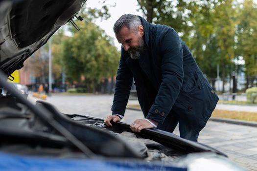 mature man trying to figure out a car breakdown leaning over the engine in the city.
