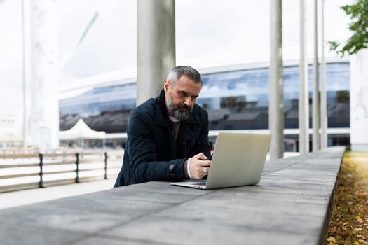 businessman man with a gray beard works on a laptop in a city park against the backdrop of an office building.