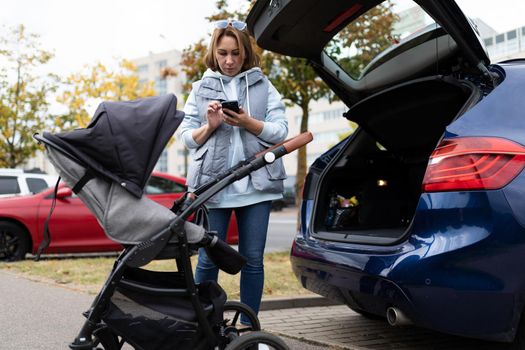 a young mother, using an application on her mobile phone, is trying to figure out how to load and fold a baby stroller into the trunk of a car.