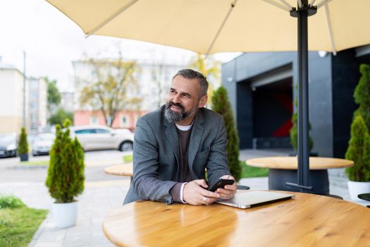Gray-haired man Businessman with a beard works in a city cafe on a laptop holding a phone in his hands.