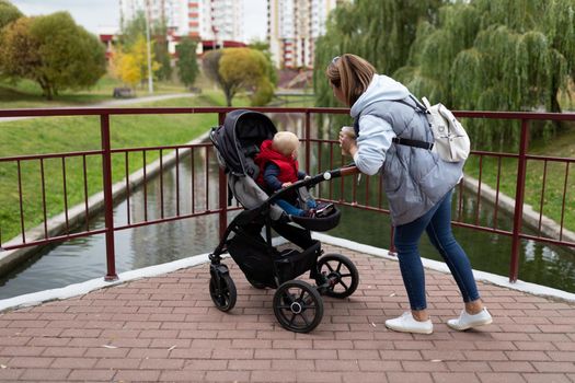 a young mother with a backpack on her shoulders walks with her child in a pram in the city park.