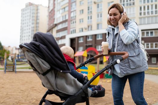 a young mother listens to music on headphones and drinks coffee while walking with a baby in a pram on a playground near the house.