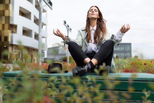 young stylish woman meditates during a lunch break in the park against the backdrop of an office building.
