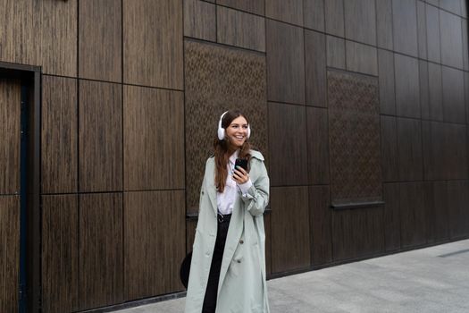 portrait of a stylish young woman in headphones using a mobile phone against the background of a brown wall of a modern building.