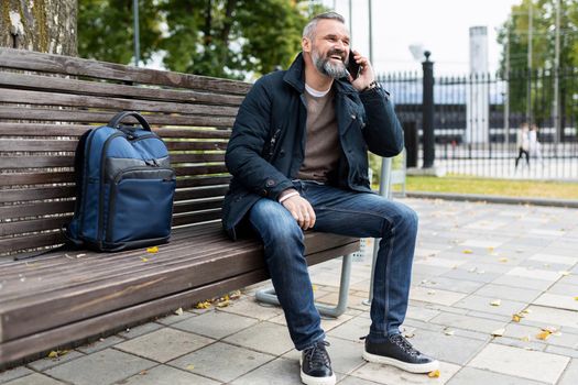 a man with a gray beard smiling speaks on the phone while walking in the park sitting on a bench.