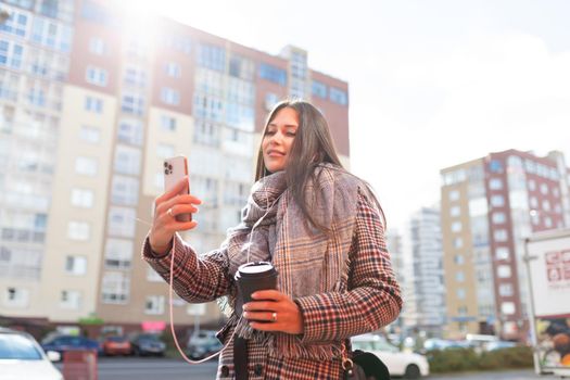 stylish young woman walking with a cup of coffee and taking a selfie in headphones.
