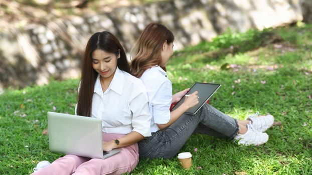 Two young woman sitting in public park and working with computer laptop.