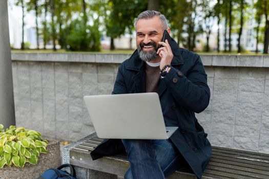 mature male entrepreneur working online on a laptop in a park in a city talking on the phone with a smile.