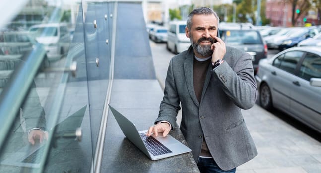 a businessman in a gray jacket works online on a laptop next to an office building and speaks on a mobile phone with a smile.