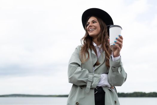 young stylish woman drinking coffee with a smile on her face looking away on the background of the embankment.