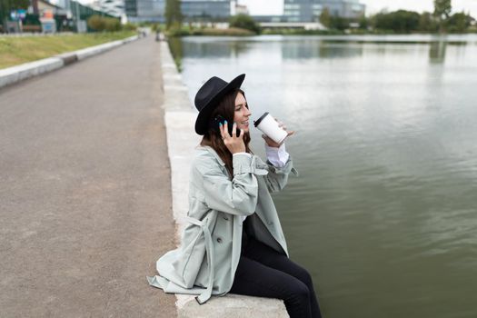 a young woman sits on the embankment dangling her legs to the water and drinking coffee talking on the phone looks into the distance.