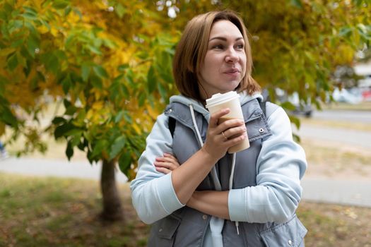 portrait of a young woman with a slight smile with a glass of coffee against the background of an autumn tree in the park.