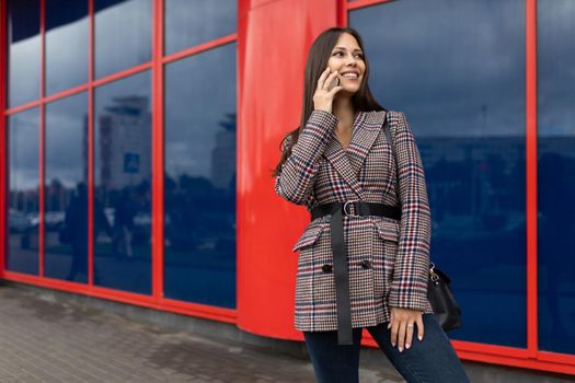 a young woman in an autumn coat speaks on a mobile phone against the background of a red blue building facade.