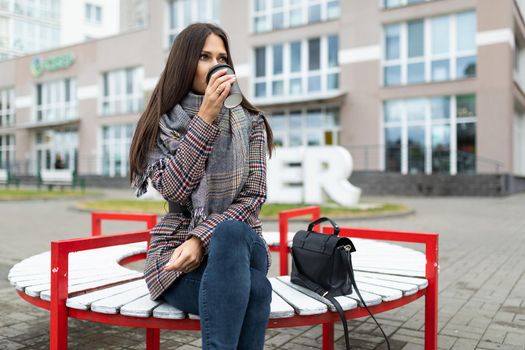a young woman in autumn clothes drinks coffee while sitting on a bench against the background of a building.