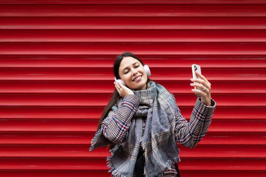 young charismatic woman dancing in headphones holding a mobile phone in her hands against the background of a bright red embossed wall.