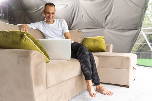 A smiling man is sitting on the comfy couch in the living room, relaxing and talking to his friend or family on the laptop, staying connected.