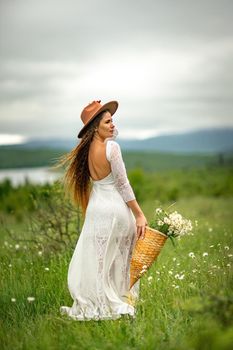 A middle-aged woman in a white dress and brown hat stands on a green field and holds a basket in her hands with a large bouquet of daisies. In the background there are mountains and a lake