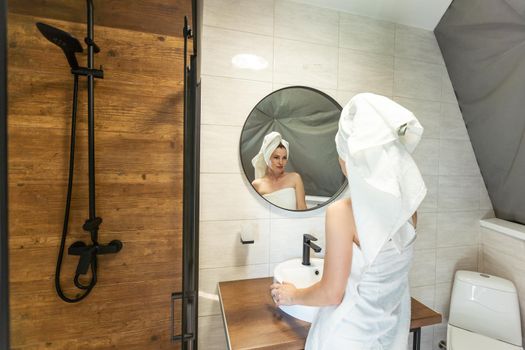 After shower body and head wrapped in towel 35s woman looks in mirror touches moisturized soft healthy face skin feels satisfied enjoy spa cosmetics treatment procedure, morning care hygiene concept