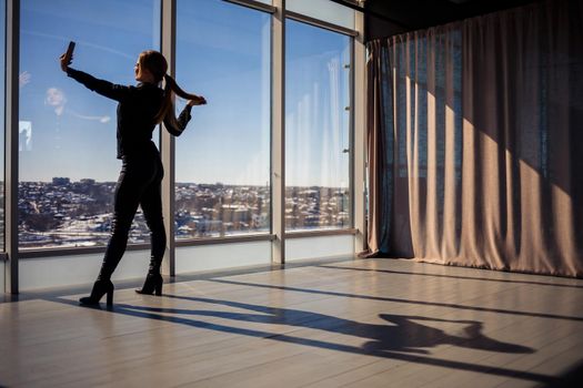 A beautiful girl takes a selfie on her phone against the background of panoramic windows in a skyscraper