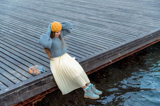 Outdoors fashion portrait of a beautiful middle aged woman walking on the beach. Marine background. Dressed in a stylish warm blue sweater, yellow skirt and beret