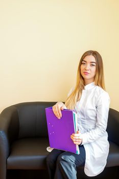 Portrait of a young caucasian doctor with folders and blonde long hair wearing a white coat sitting on the black sofa looking at the camera