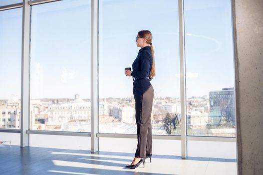 Confident business woman boss standing in modern office or hotel window enjoying big city view, woman leader, business owner thinking about future success, planning new opportunities,