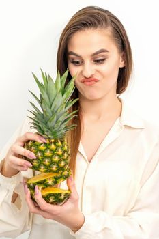 Beautiful young Caucasian woman holding pineapple and smiling, wearing a white shirt over white background