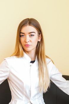 Portrait of a young caucasian doctor with blonde long hair wearing a white coat sitting on the black sofa looking at the camera