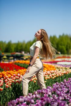 Beautiful woman in hat laughing to camera in spring flower garden. Girl with camera smiling in park with many flowers. Woman enjoying time in blooming flowers field. Pretty lady walking in meadow.