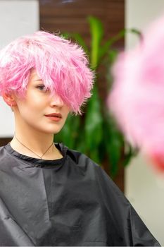 The beautiful young caucasian woman with a new pink short hairstyle in the mirror reflection looking at the camera in a hairdresser salon