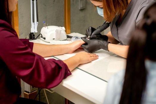 Manicure master wearing protective mask uses electric nail file machine in a nail salon, close up