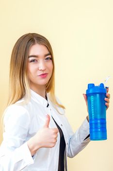 The young Caucasian female doctor wearing a white coat holds a blue plastic bottle with water with thumb up looking at the camera