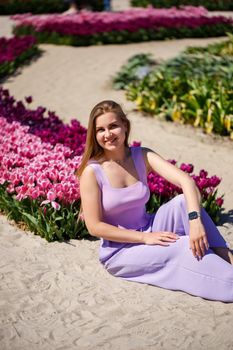 Back view of anonymous woman in summer dress and hat walking near blooming tulips in meadow on sunny day in nature