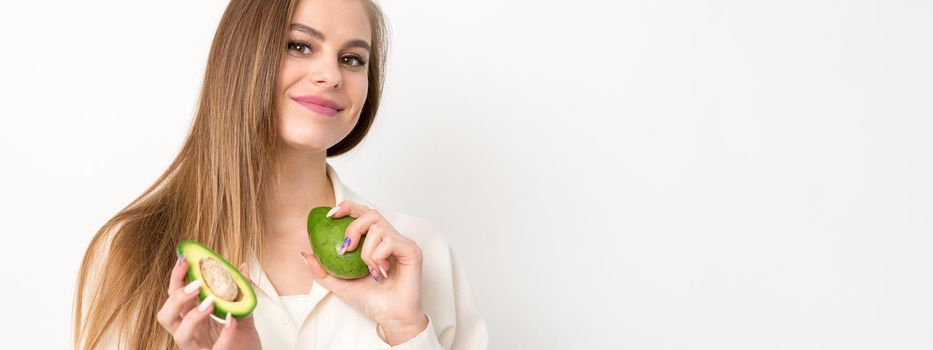 Portrait of a lovely smiling young brunette caucasian woman wearing the white shirt with long hair holding and showing avocado, standing isolated over white background