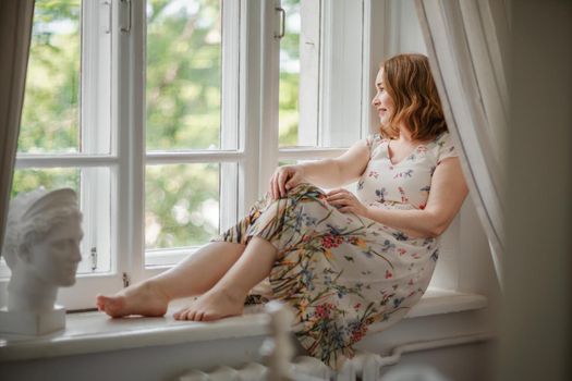 A middle-aged woman in a cream dress sits mysteriously and looks out the window on the windowsill. Green trees outside