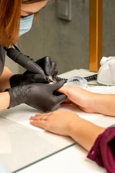 Manicure master wearing protective mask uses electric nail file machine in a nail salon, close up