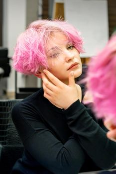 The beautiful young caucasian woman with a new short pink hairstyle looking at her reflection in the mirror checking hairstyle in a hairdresser salon