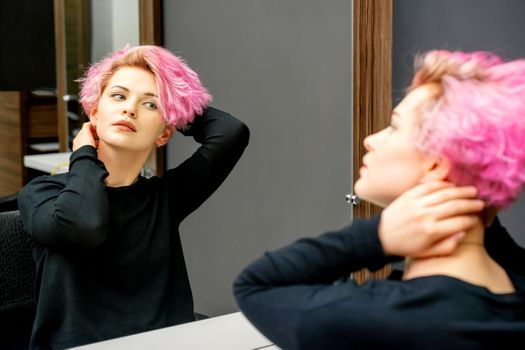 The beautiful young caucasian woman with a new short pink hairstyle looking at her reflection in the mirror checking hairstyle in a hairdresser salon
