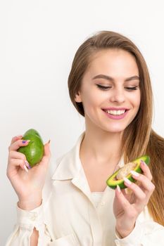 Portrait of a lovely smiling young brunette caucasian woman wearing the white shirt with long hair holding and showing avocado, standing isolated over white background