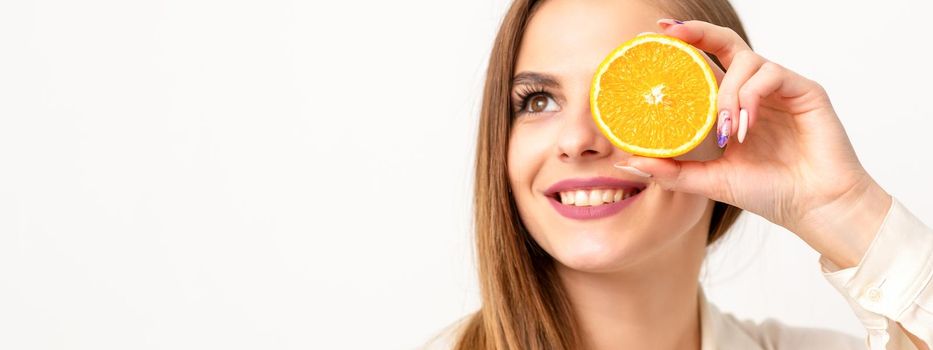 Portrait of a cheerful caucasian young woman covering eye with an orange slice wearing a white shirt over a white background