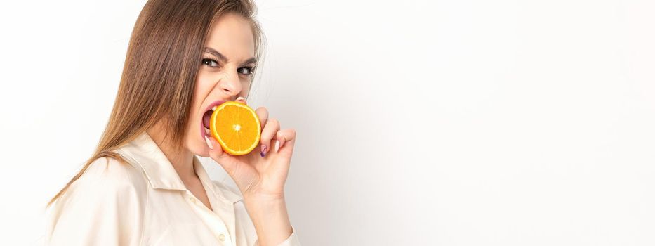 Young caucasian pretty cunning brunette woman biting one orange half and looking at the camera wearing a white shirt over white background