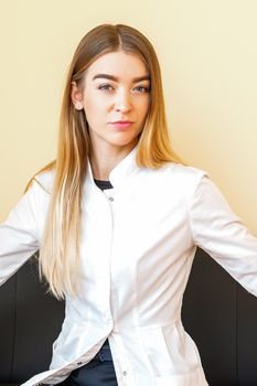 Portrait of a young caucasian doctor with blonde long hair wearing a white coat sitting on the black sofa looking at the camera