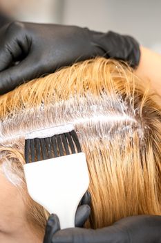 The hairdresser is dyeing blonde hair roots with a brush for a young woman in a hair salon