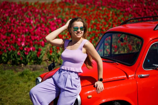 A girl model in casual clothes sits on a red car among tulip fields.