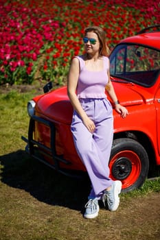 A girl model in casual clothes sits on a red car among tulip fields.